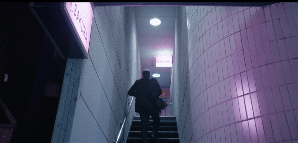 A figure in a blue blazer ascends steps in a white concrete building with a pink florescent signage reading "sakura" in the documentary Snack Sakura.